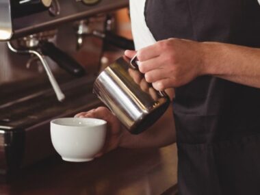 Barista v černé zástěře, zaměřený na důvody pro zdokonalování svého řemesla, nalévá mléko do bílého šálku kávy u pultu plného zařízení na přípravu kávy.